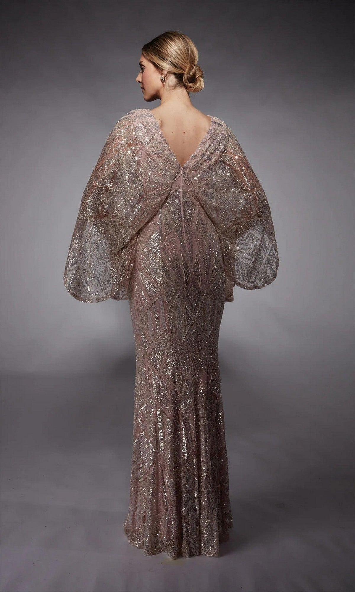A woman with blonde hair in a low bun models the Alyce 27756 long formal dress, featuring floor-length geometric sequins and wide, sheer sleeves, as she stands with her back to the camera against a plain gray backdrop.